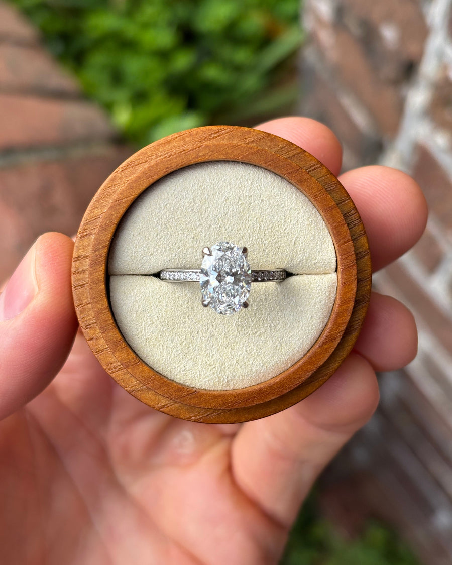 Oval lab diamond solitaire engagement ring with a pavé band set in platinum inside a wooden ring box.