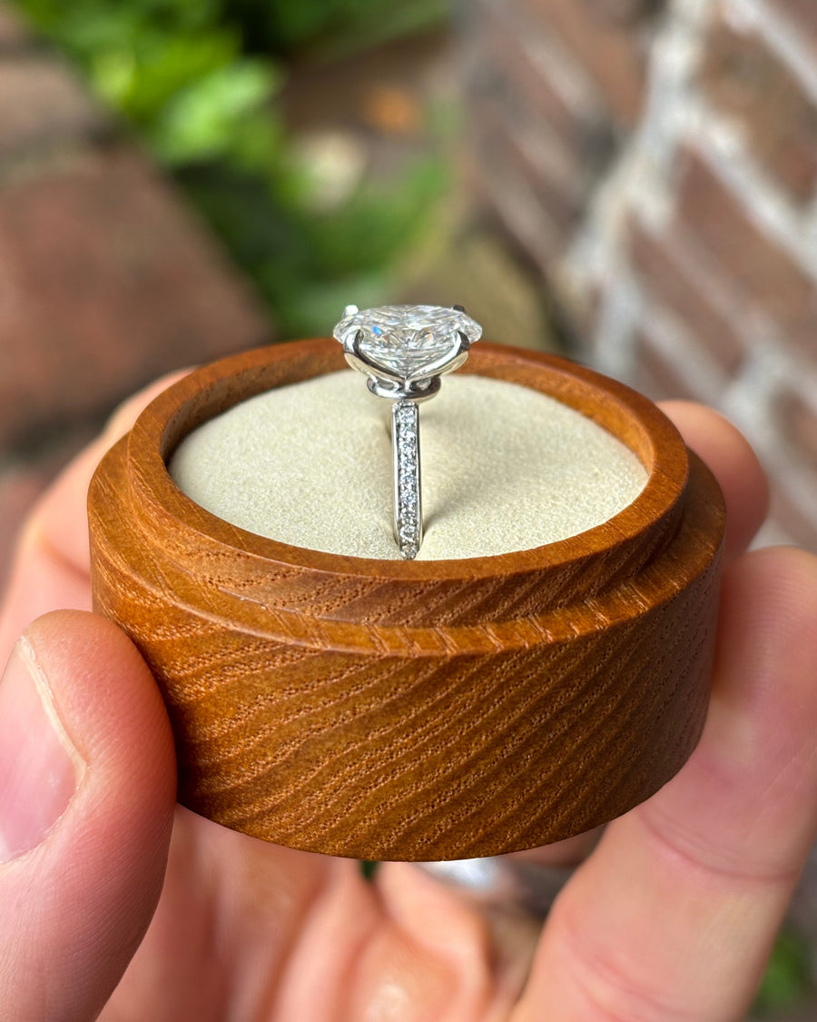 Oval lab diamond solitaire engagement ring with a pavé band set in white metal inside a wooden box.