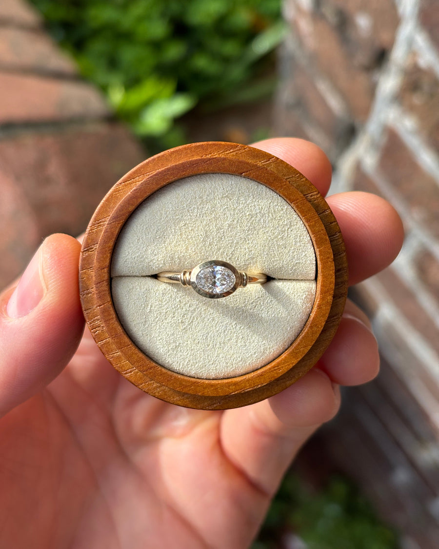 0.7ct oval diamond engagement ring in a yellow gold east-west bezel setting held in a wooden ring box.