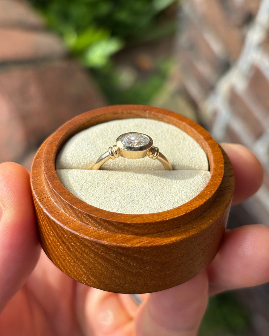 Oval diamond engagement ring in a gold bezel setting, held in a small wooden ring box.