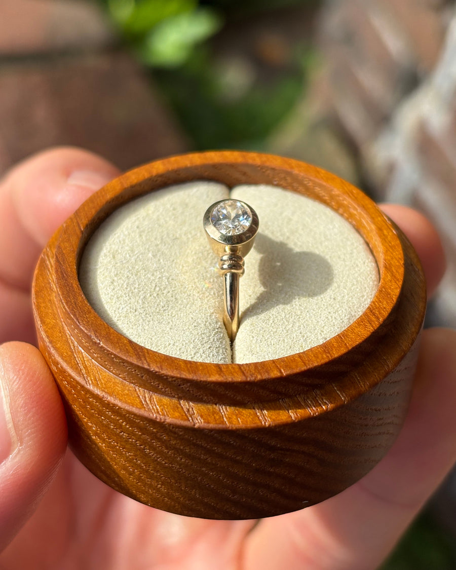 Gold bezel-set diamond solitaire ring in a round wooden box.