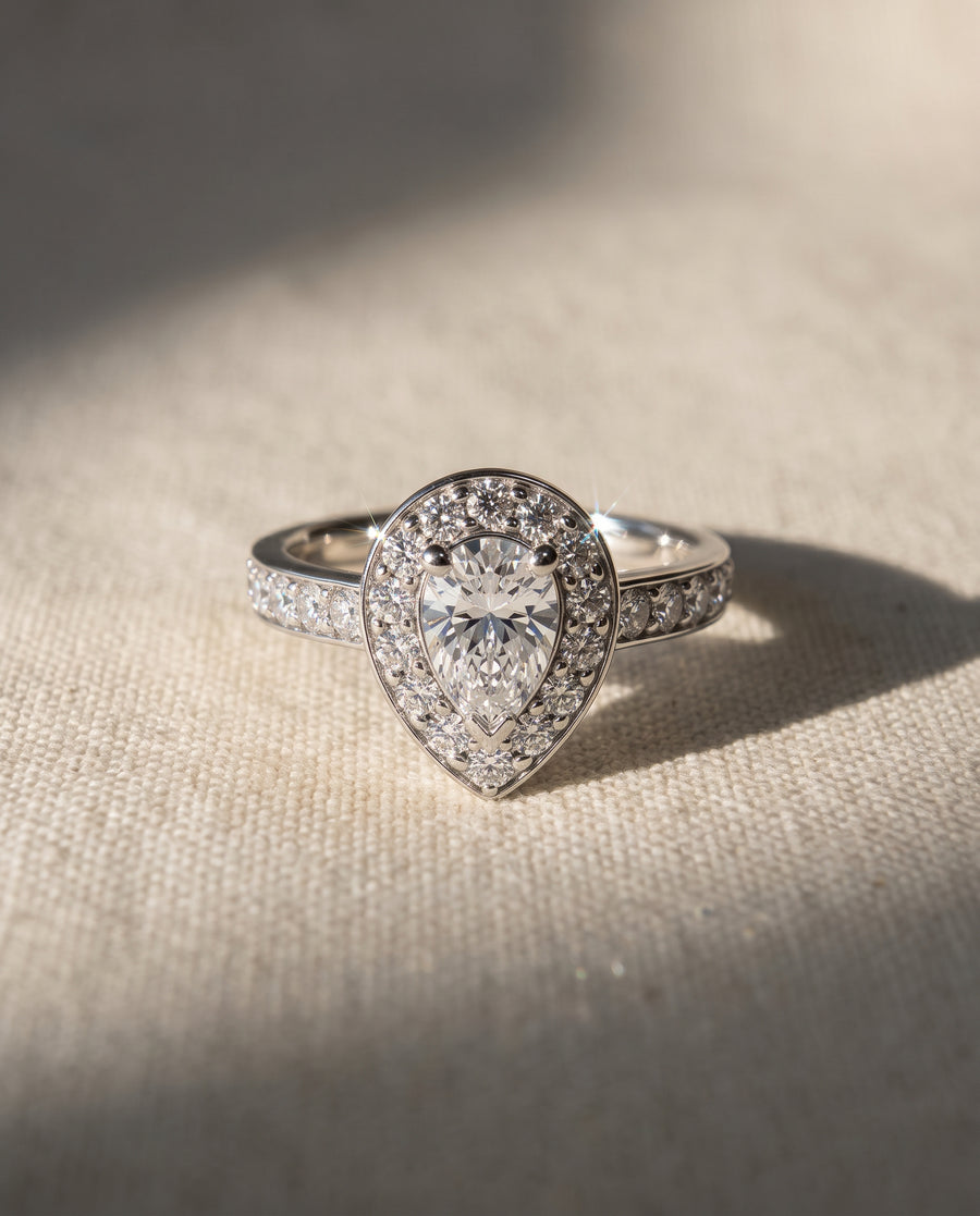 Pear-shaped diamond cluster engagement ring in polished silver-tone metal with a halo setting on a textured surface.