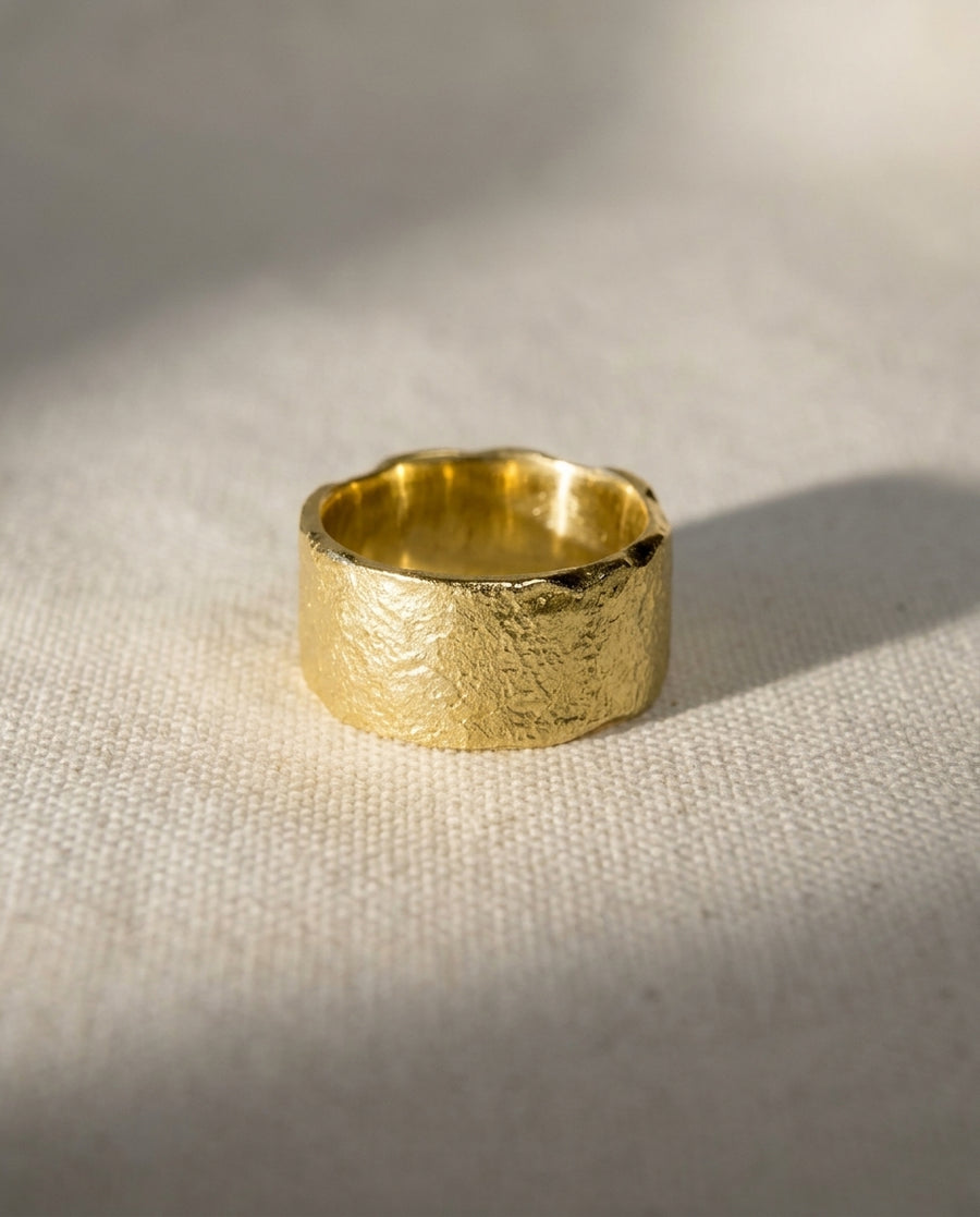 Wide gold wedding band with a textured, reticulated surface sitting on a neutral fabric background.