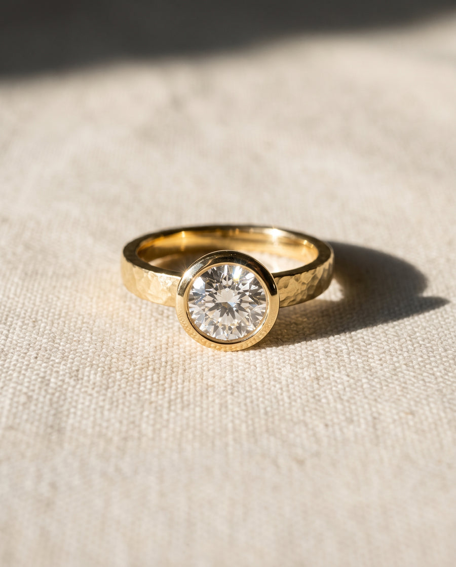 Gold engagement ring with a round brilliant cut diamond in a hammered bezel setting on textured fabric.