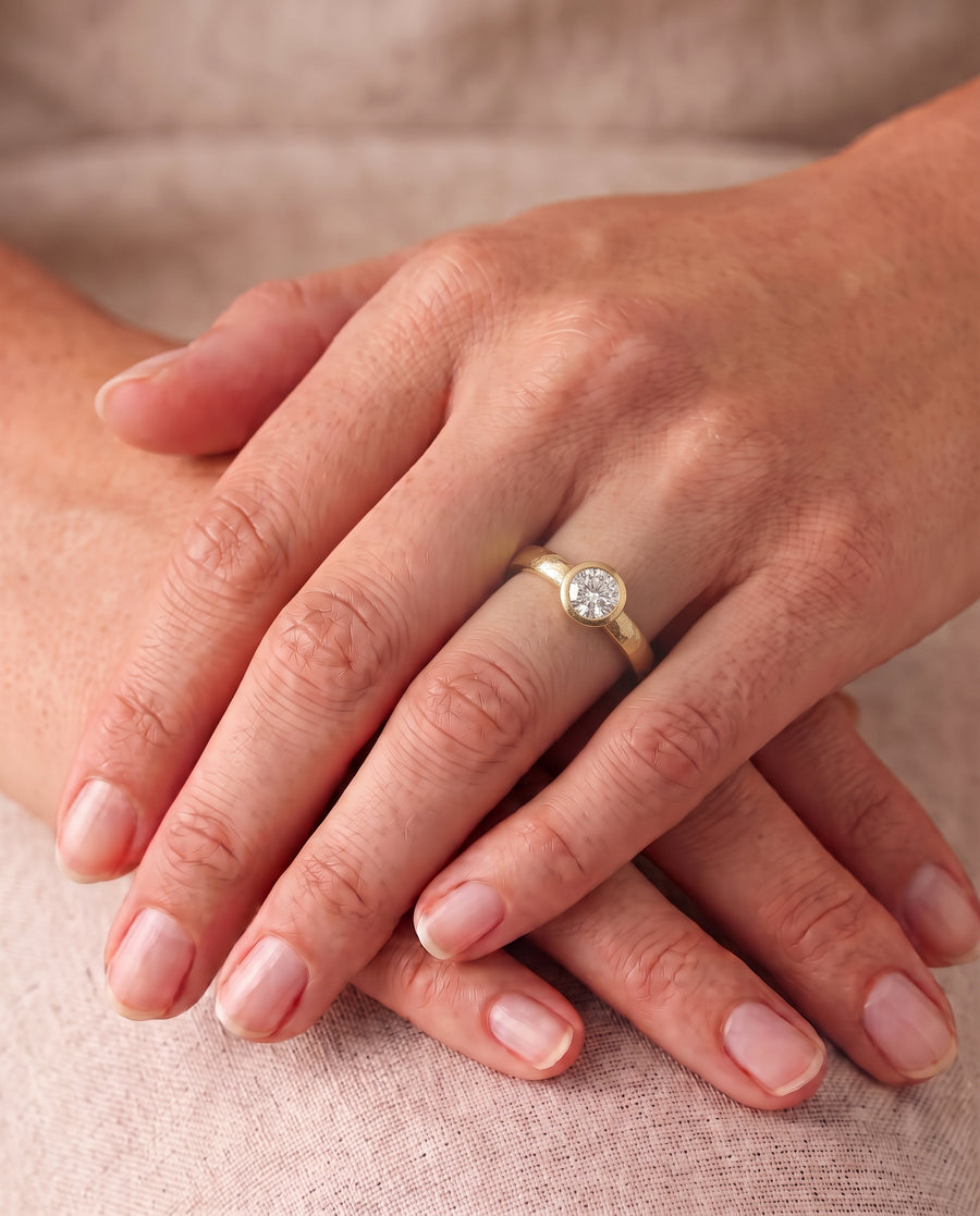 Round brilliant cut diamond engagement ring with a hammered gold bezel setting on a finger.