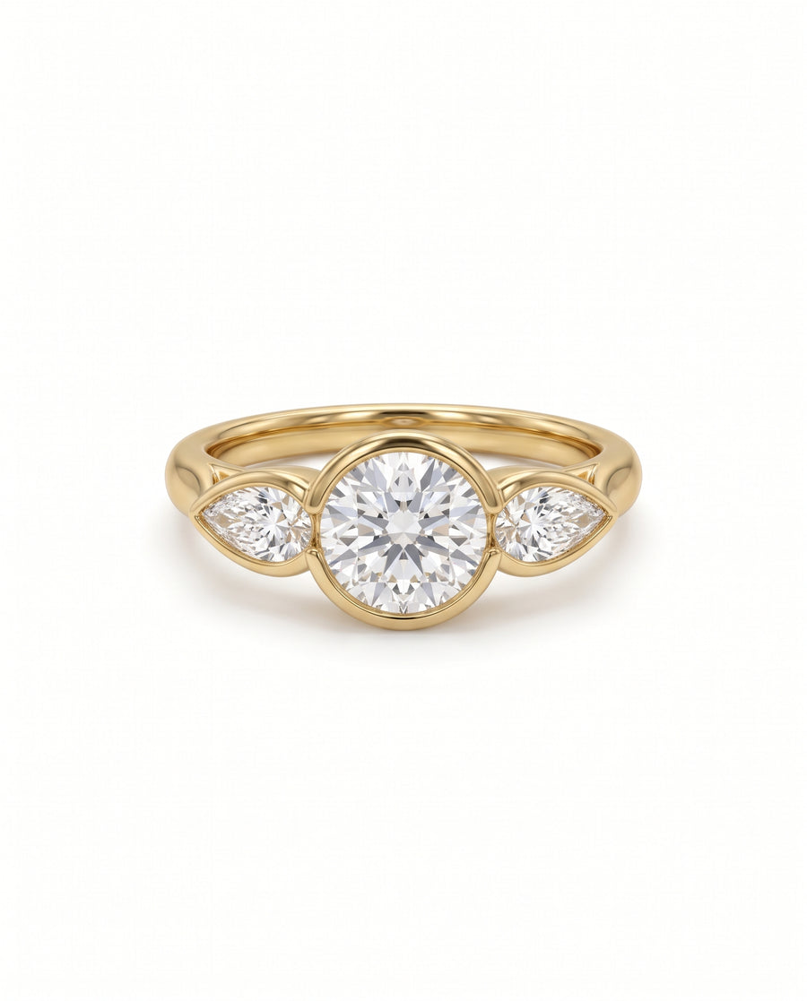 Gold trilogy engagement ring with a round brilliant center diamond and two pear shaped side diamonds in bezel settings.