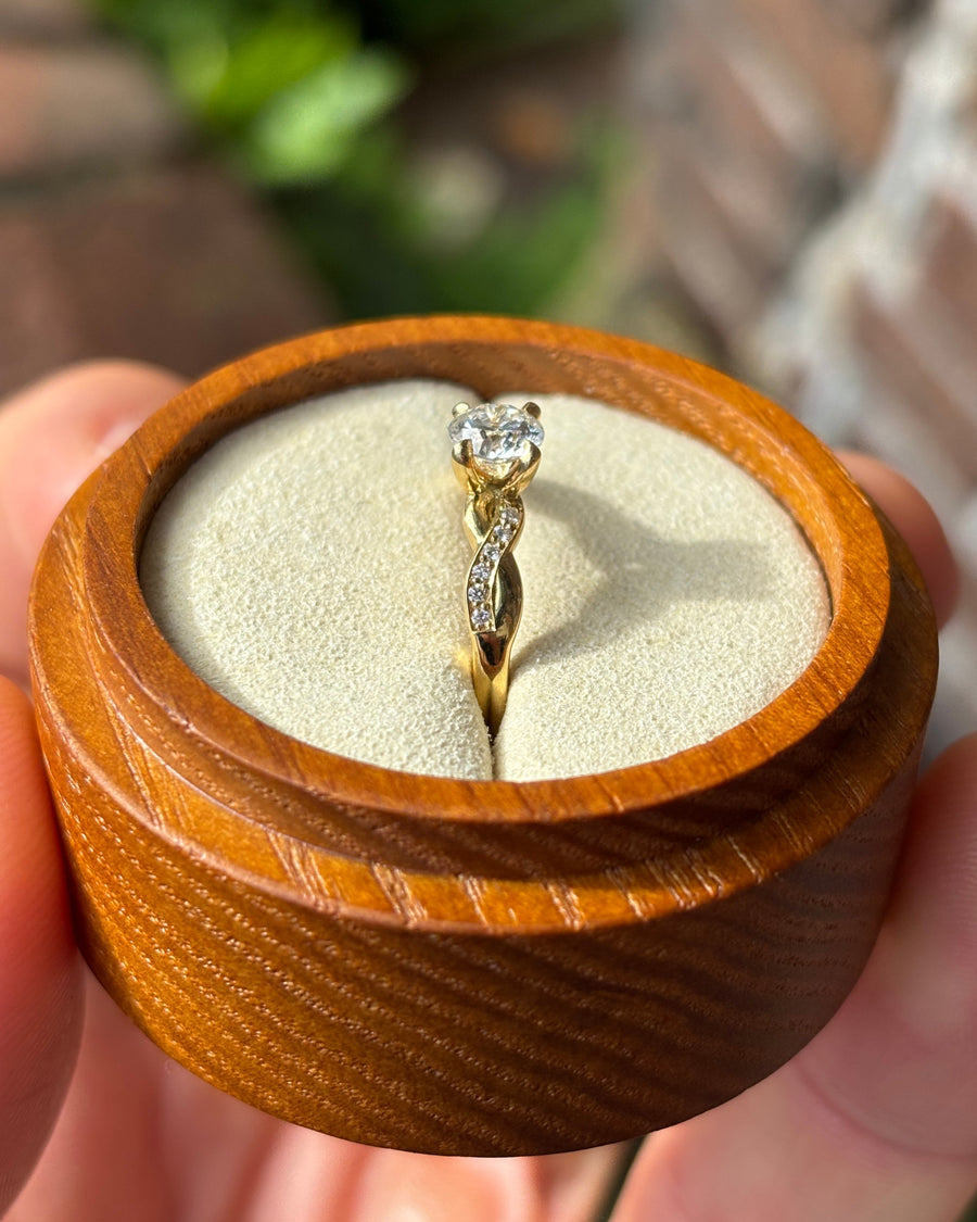 Round cut diamond engagement ring with a gold twist band in a wooden ring box.