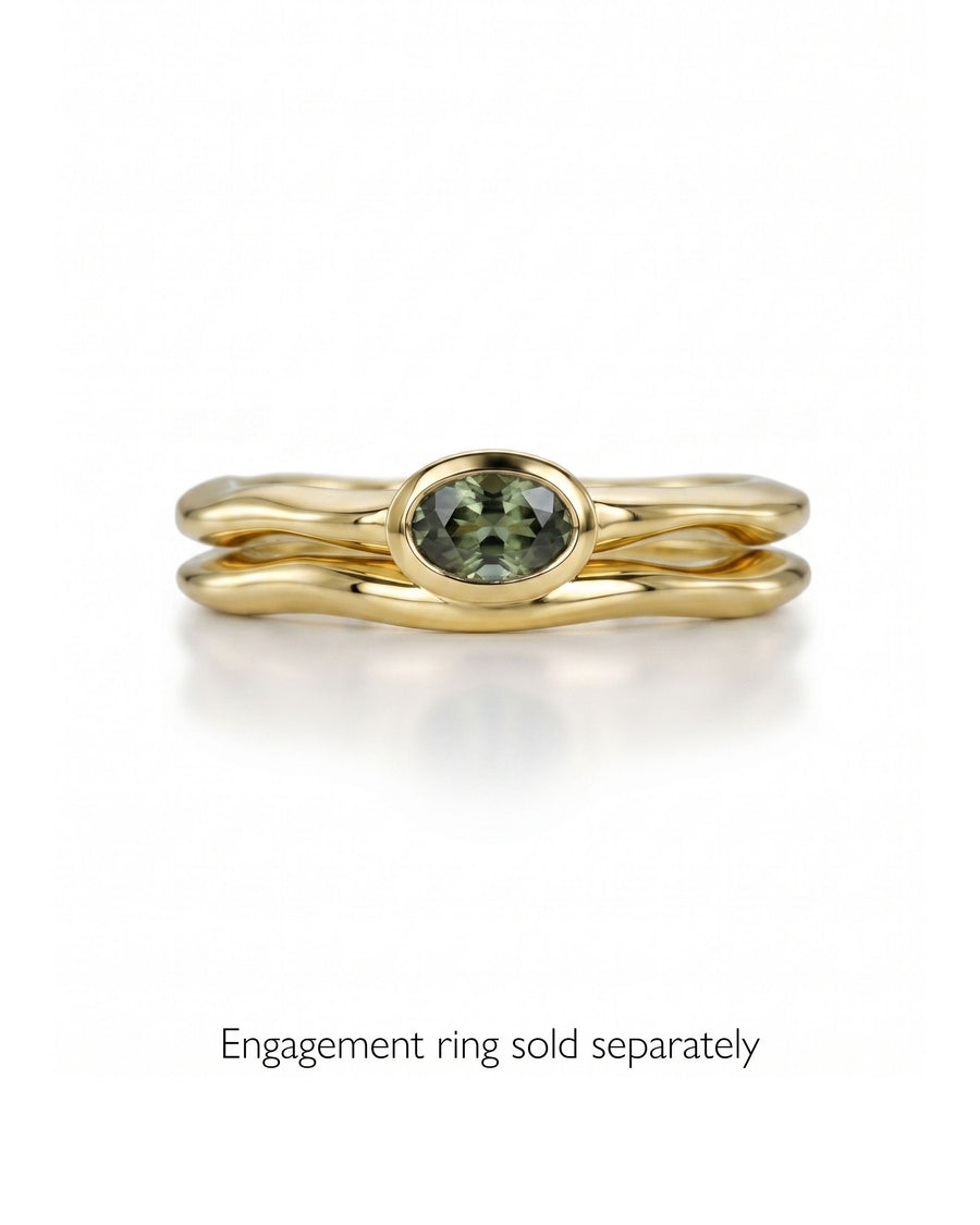 Dainty gold wedding ring with an uneven, wavy band design and a bezel-set oval green gemstone.