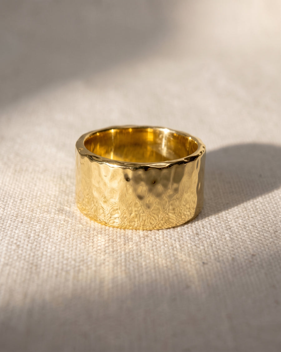 Wide gold wedding band with a textured, hammered finish resting on a light fabric surface.