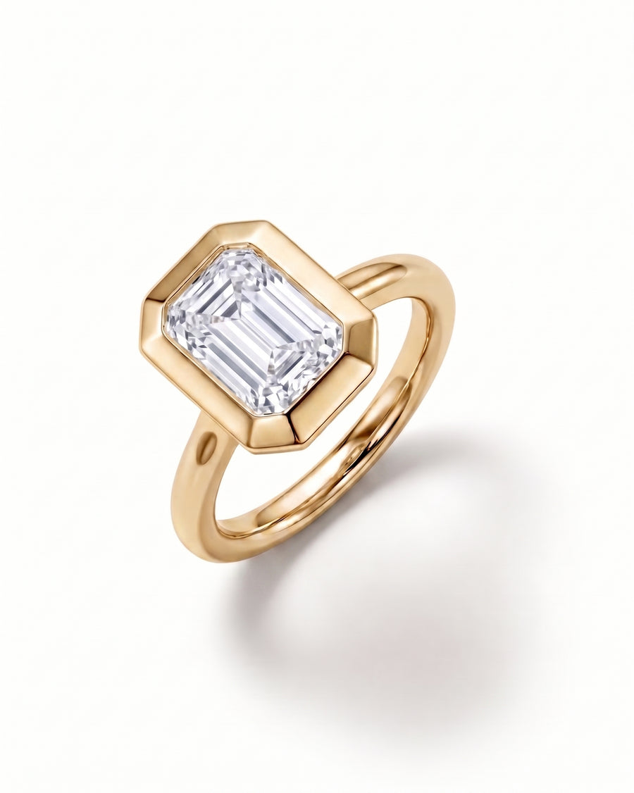 A yellow gold ring featuring a large, faceted bezel-set emerald-cut clear gemstone, shown on a clean white background with a soft shadow.