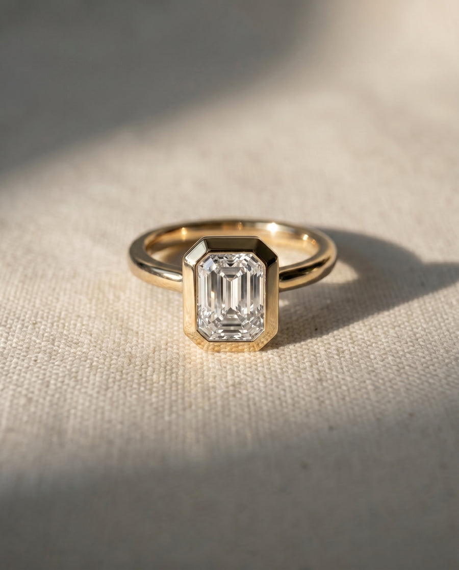 A yellow gold ring featuring an emerald-cut clear gemstone in a geometric bezel setting, resting on a textured beige linen surface with soft diagonal shadows.