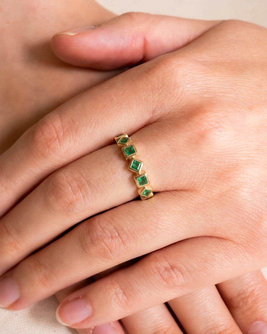 Gold eternity ring with alternating square emeralds on a hand.