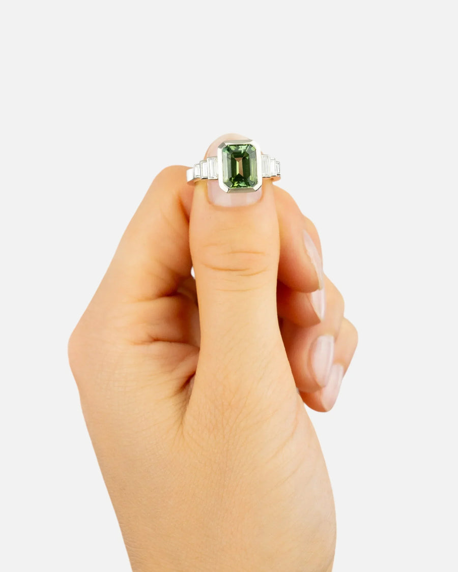 Hand holding an emerald-cut green gemstone ring with silver band against a white background