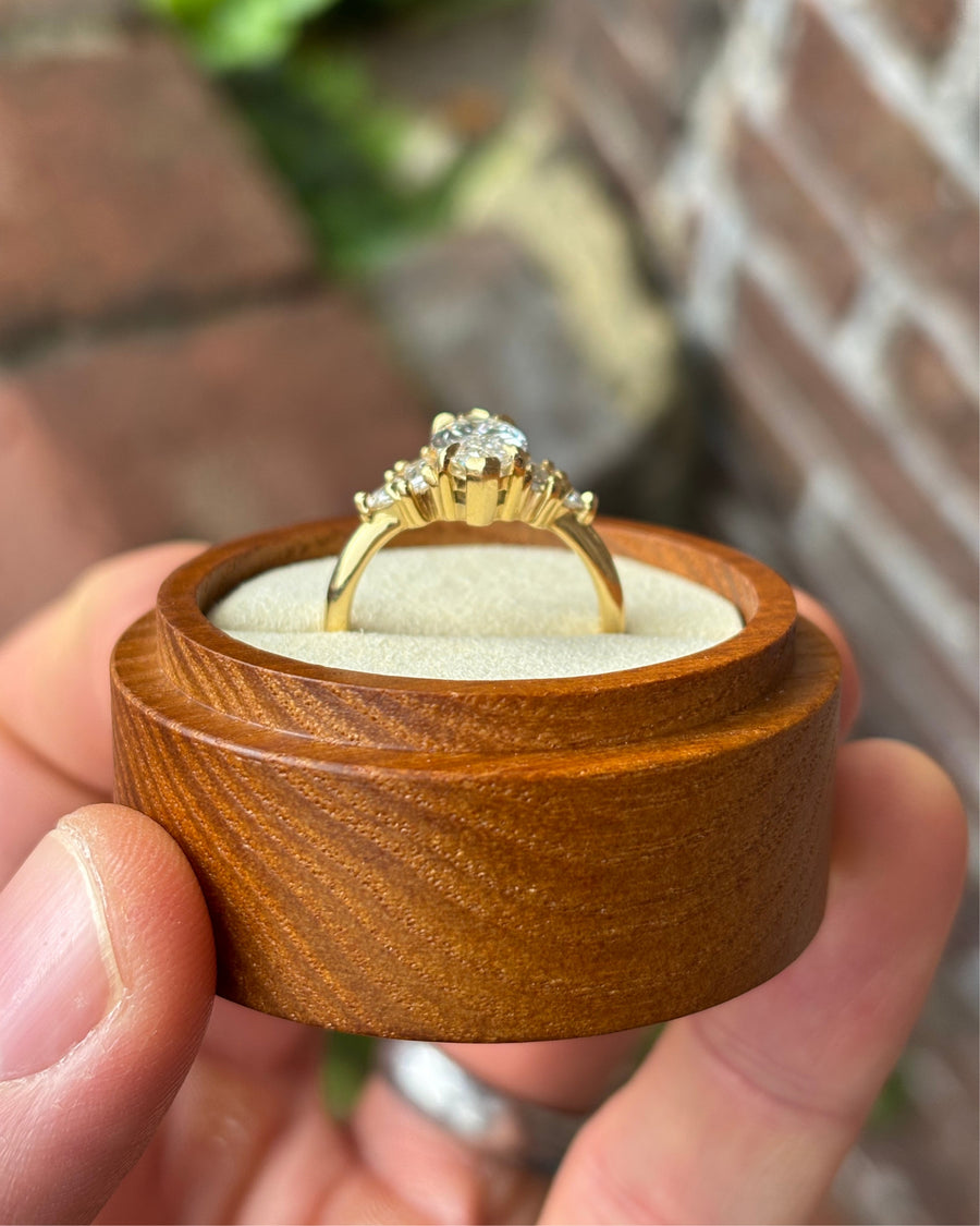 Gold engagement ring with a marquise cut center stone and claw set side diamonds in a wooden box.