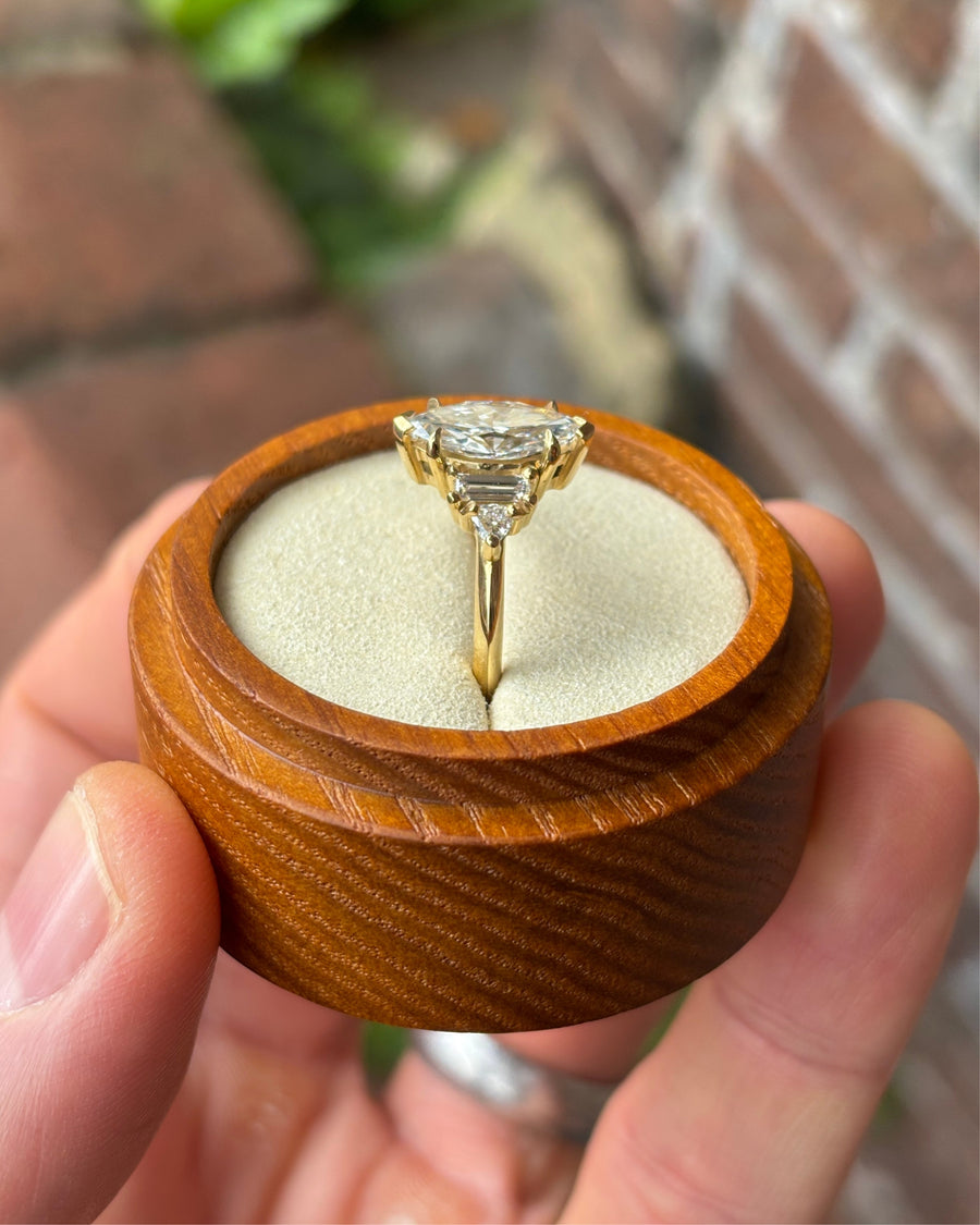 Marquise cut diamond engagement ring with side stones set in a yellow gold band inside a wooden box.