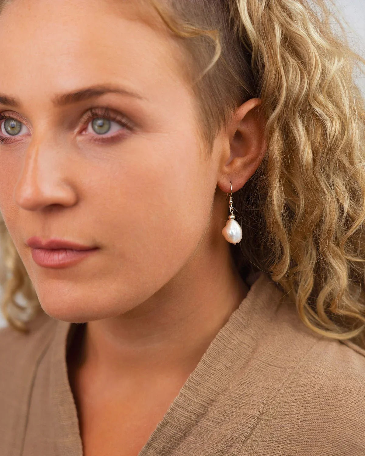 White baroque pearl drop earrings with silver hooks worn by a woman with curly blonde hair.