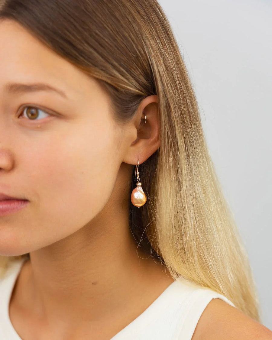 Irregular peach-colored baroque pearl drop earrings on gold hooks worn by a woman.