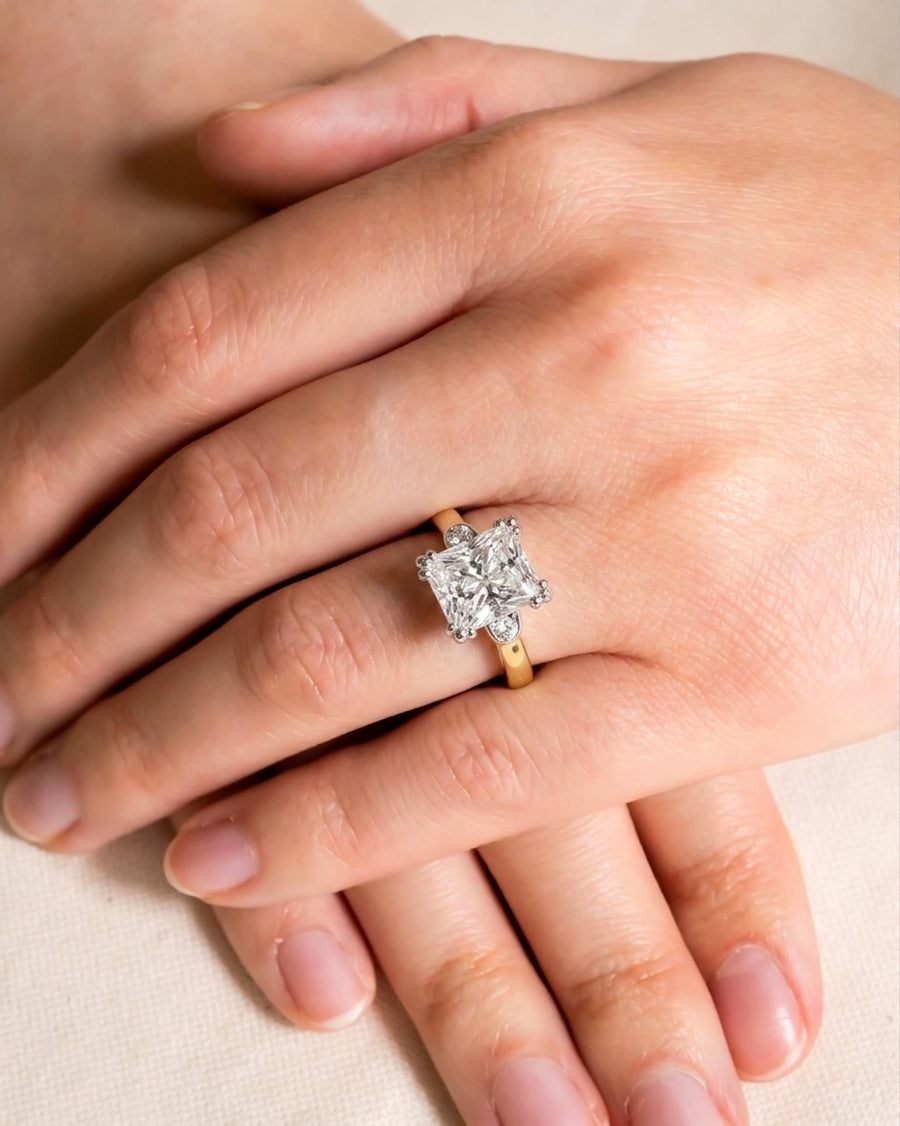 Gold engagement ring with a large princess-cut diamond and smaller side diamonds on a hand.