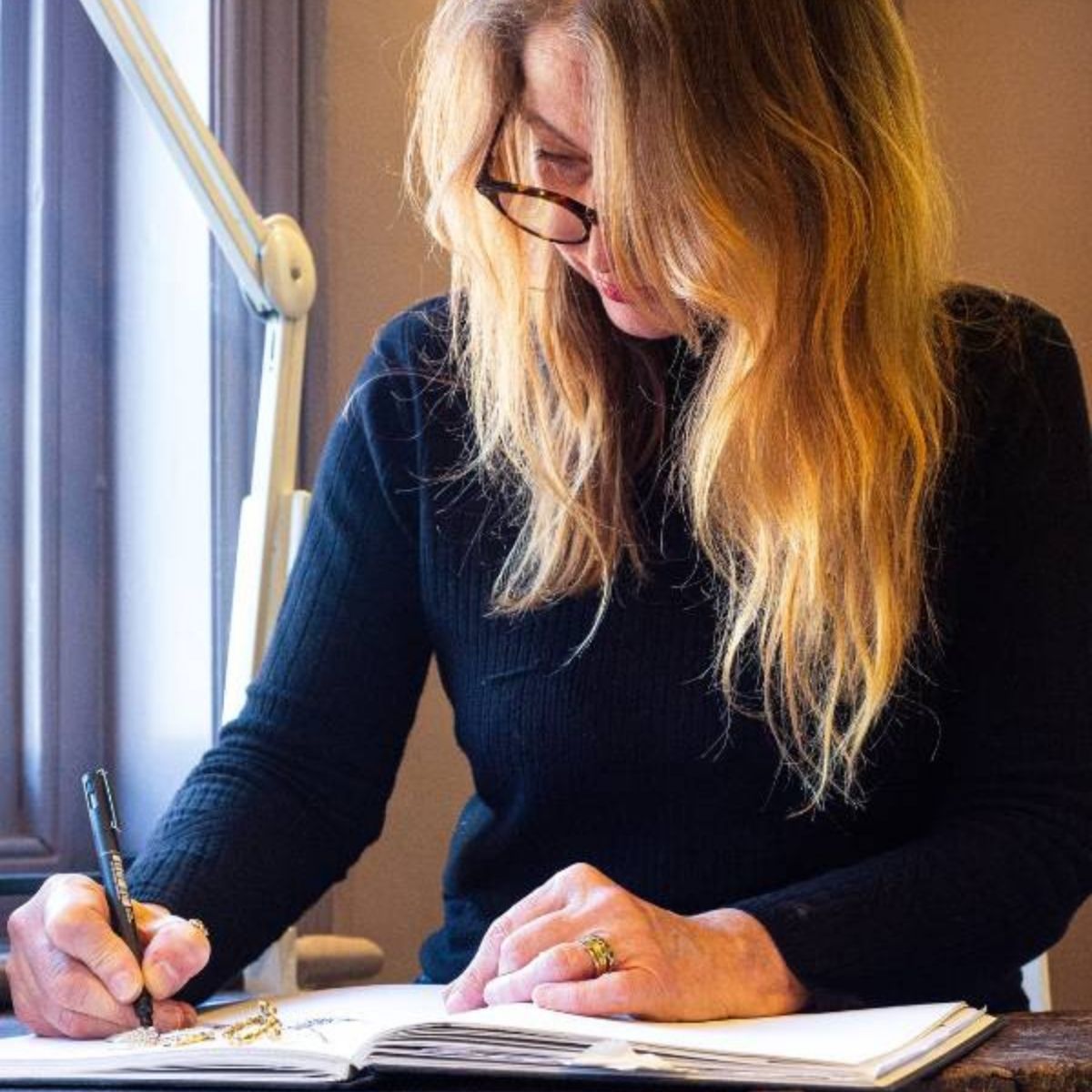 A jewellery designer sketching ideas in a notebook at a desk by a window.