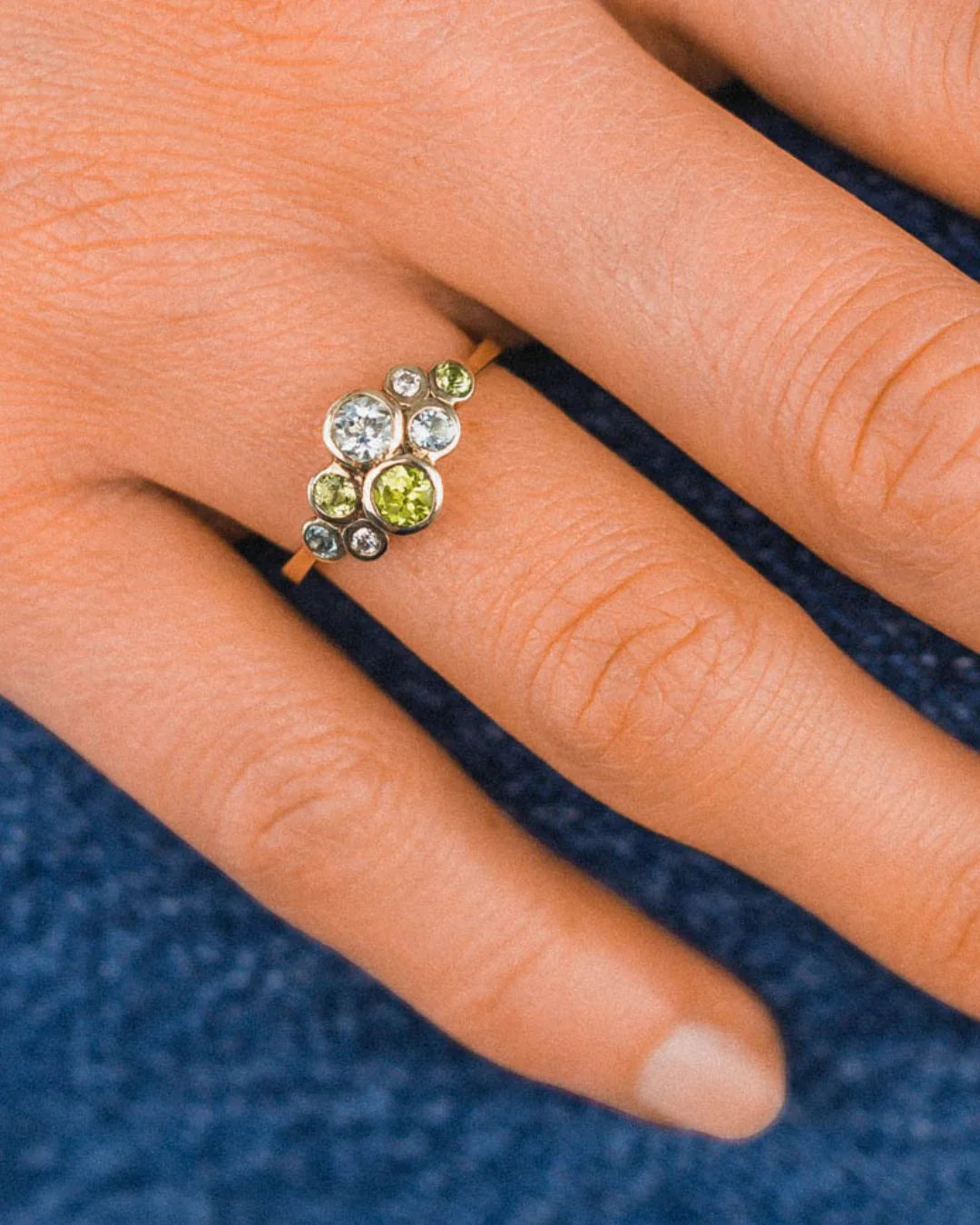 Cluster bubble ring with mixed gemstones set in gold, worn on a hand, against a blue background.