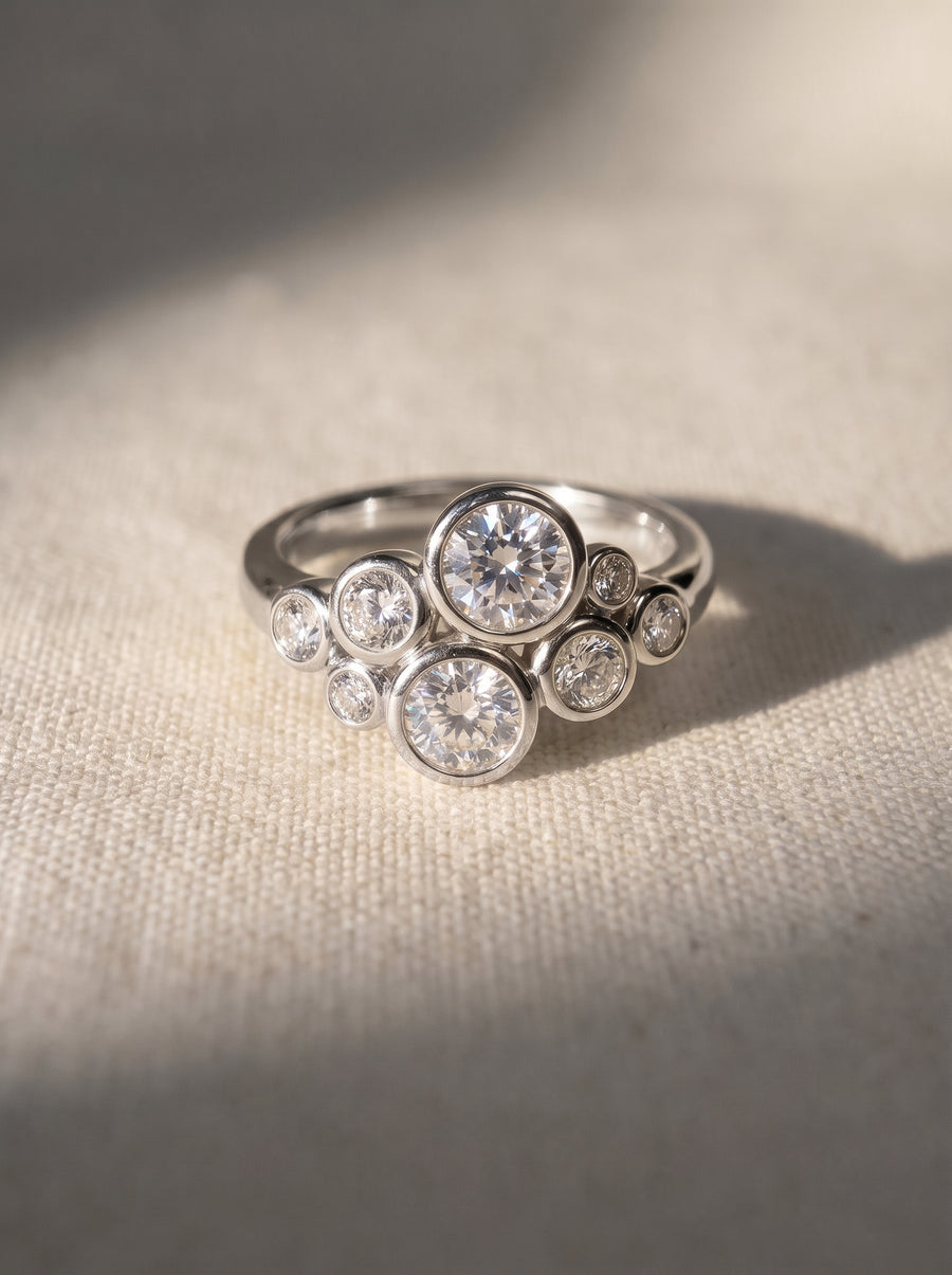 Silver cluster ring featuring round diamonds in bezel settings on a textured fabric background.
