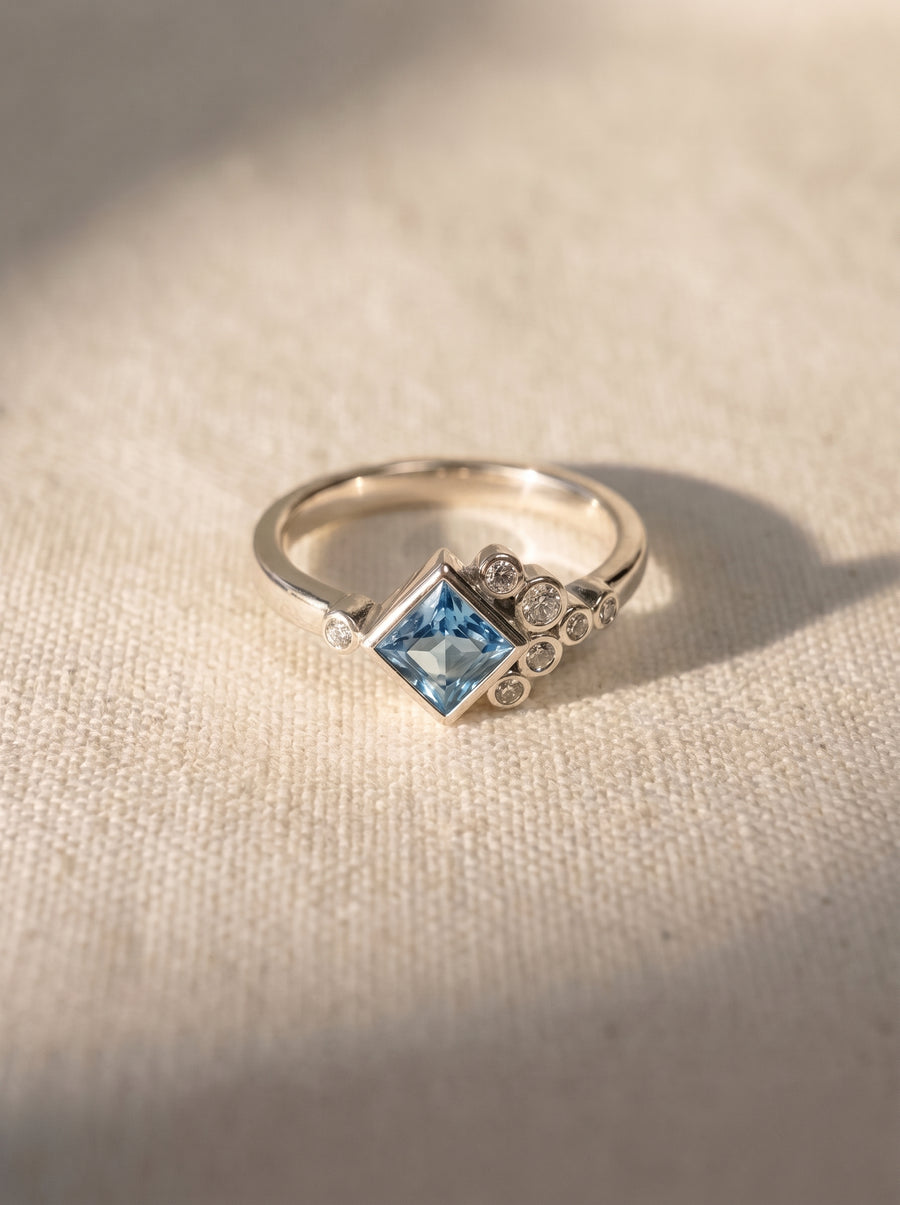 Silver ring with a square aquamarine center stone and small round diamond accents on a textured surface.