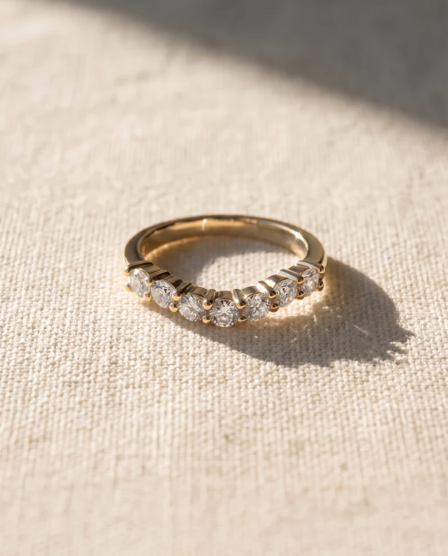 Gold wedding ring with seven round cut diamonds in a claw setting on a textured beige surface.