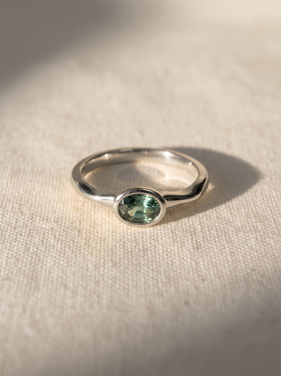Oval green sapphire engagement ring in a silver bezel setting on a textured fabric background.