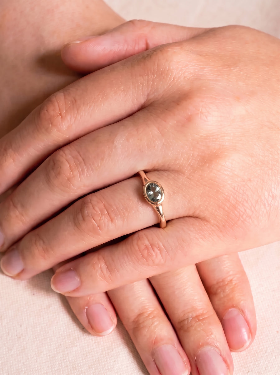 Oval sapphire engagement ring in a rose gold bezel setting worn on a finger.