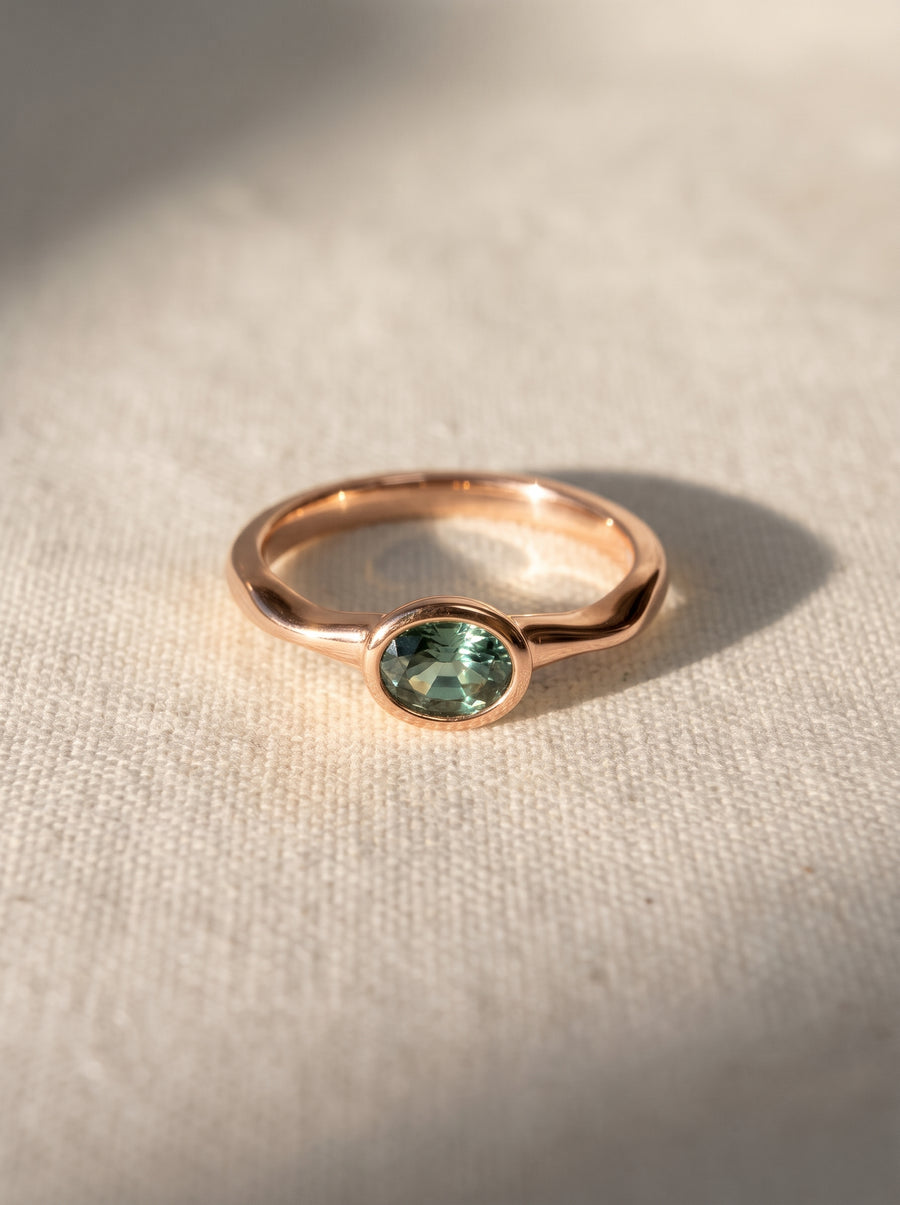 Rose gold engagement ring featuring an oval-cut green sapphire in a bezel setting on a textured fabric background.