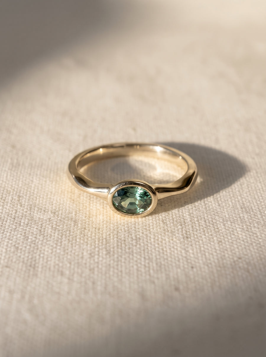 Oval green sapphire bezel set in a polished yellow gold band on a textured fabric background.