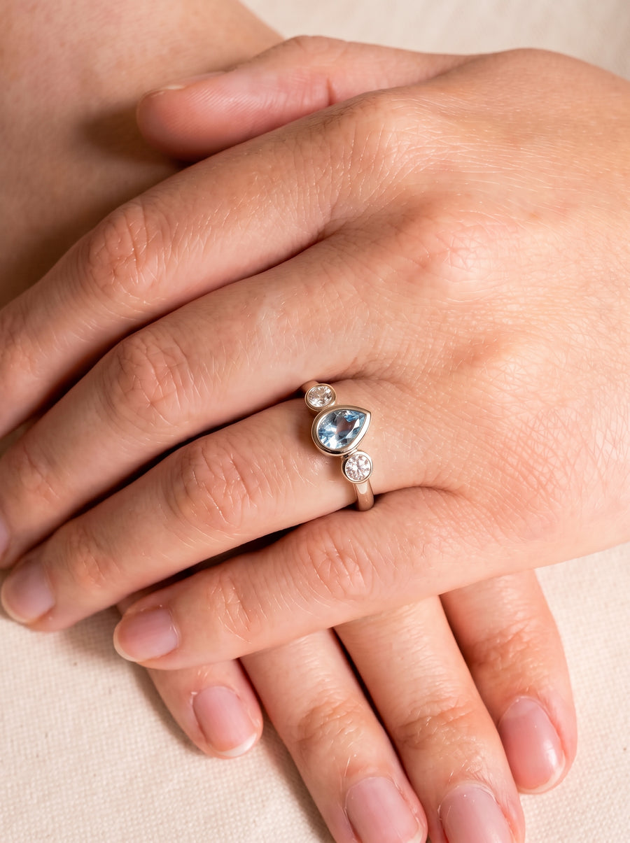 Gold trilogy ring featuring a pear-cut aquamarine flanked by two round diamonds on a finger.