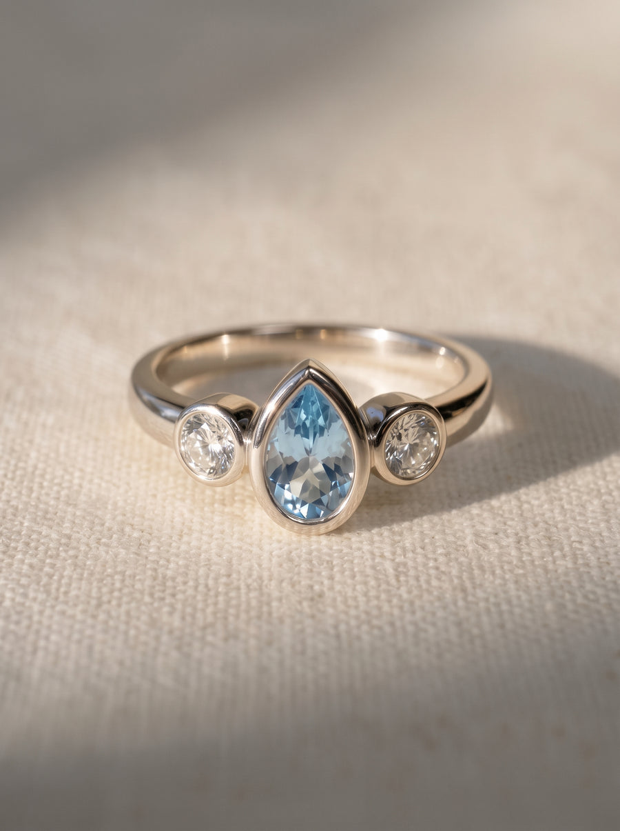 Trilogy ring in silver metal featuring a pear-cut aquamarine flanked by two round diamonds in bezel settings.