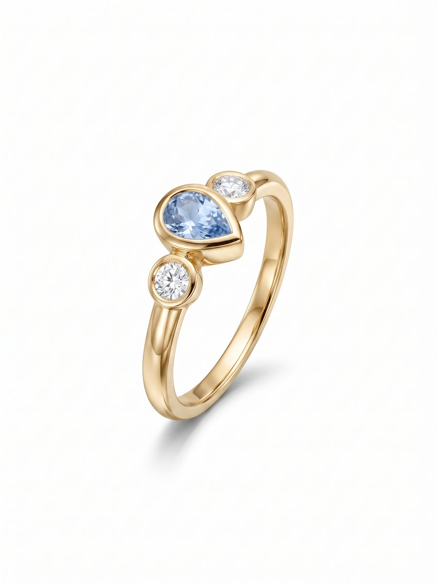 Gold trilogy ring featuring a pear-cut aquamarine flanked by two round diamonds in bezel settings.