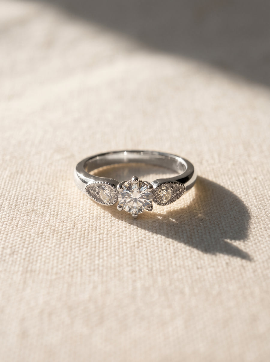 Silver engagement ring with a round center diamond and two pear-shaped side stones on a textured fabric background.