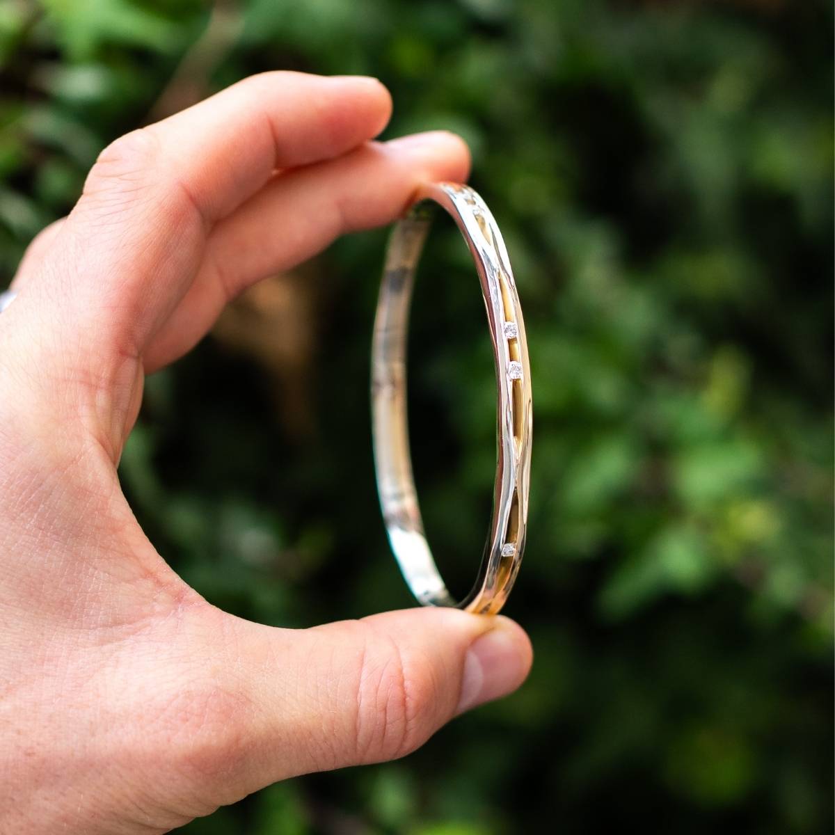 A slim silver bangle with subtle diamond details, held between fingers against a green background.