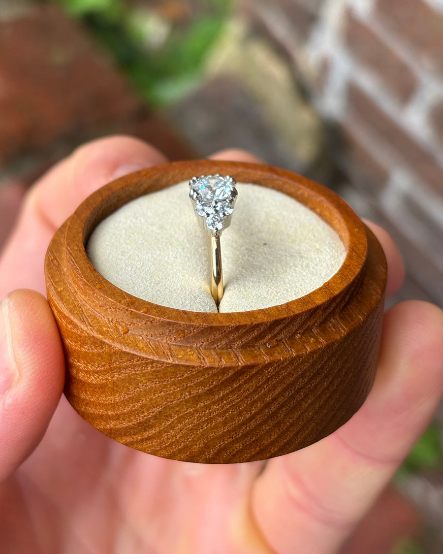 Round cut diamond engagement ring with a trefoil claw setting on a gold band in a wooden box.
