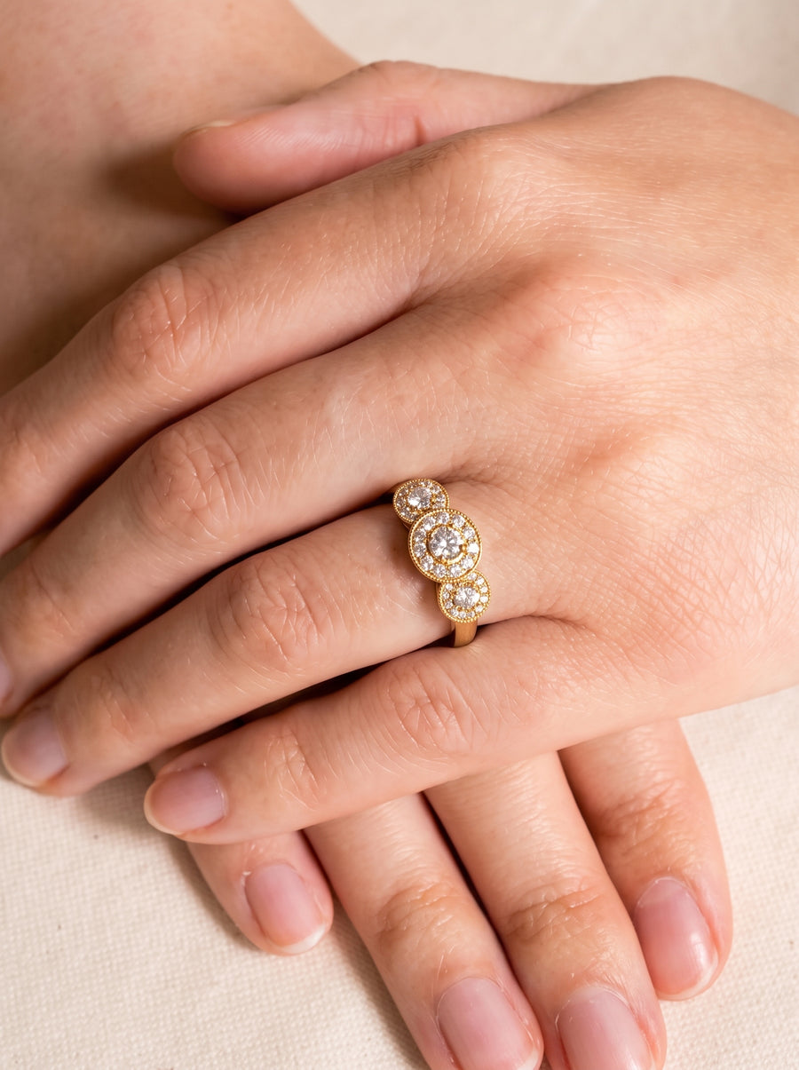Gold trilogy engagement ring featuring three round diamond cluster settings on a finger.