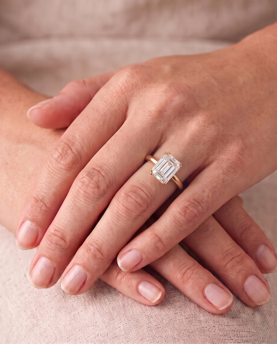 Emerald-cut diamond solitaire engagement ring with a gold cathedral band worn on a finger.