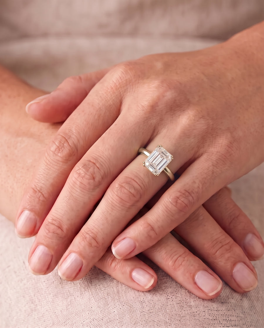 Emerald-cut diamond solitaire engagement ring with a gold cathedral band worn on a finger.