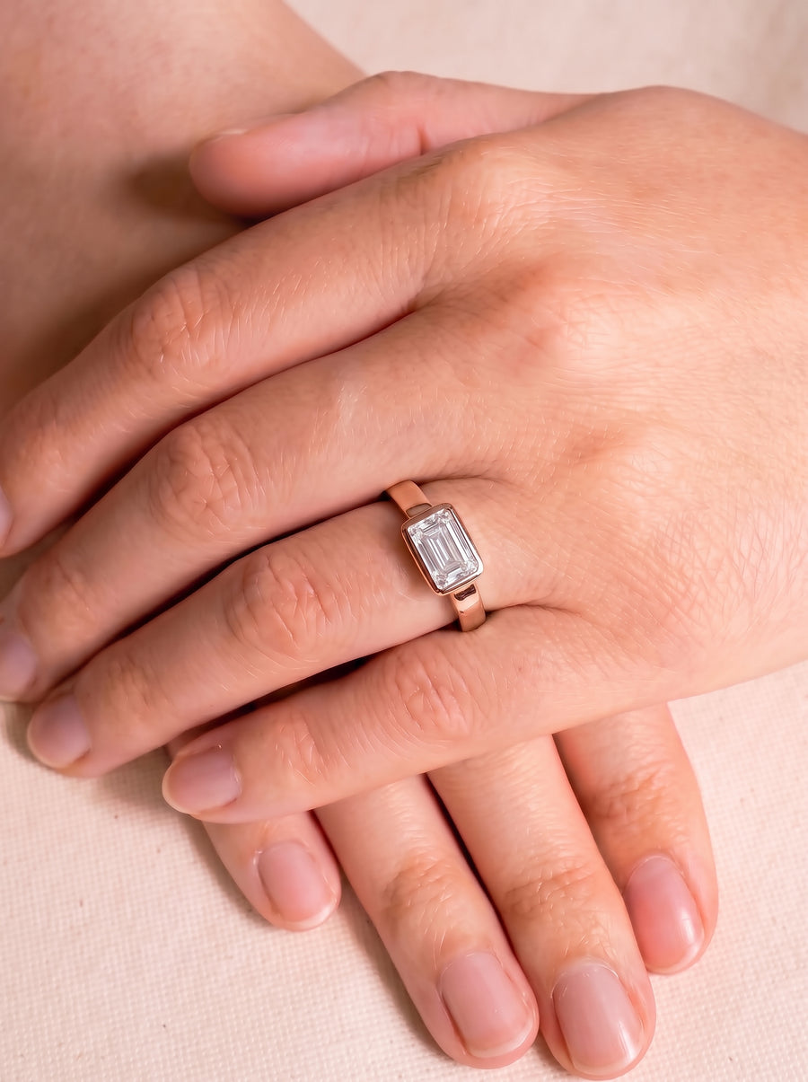 Emerald cut diamond ring in a rose gold bezel setting worn on a finger.