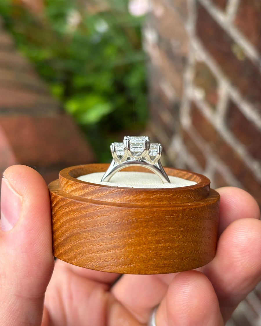 Platinum trilogy engagement ring with emerald cut diamonds and a hidden halo in a wooden ring box.