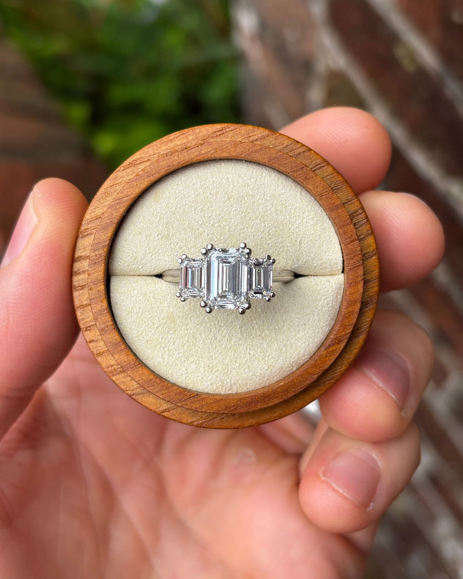 Trilogy engagement ring with emerald cut diamonds in a platinum setting inside a wooden ring box.