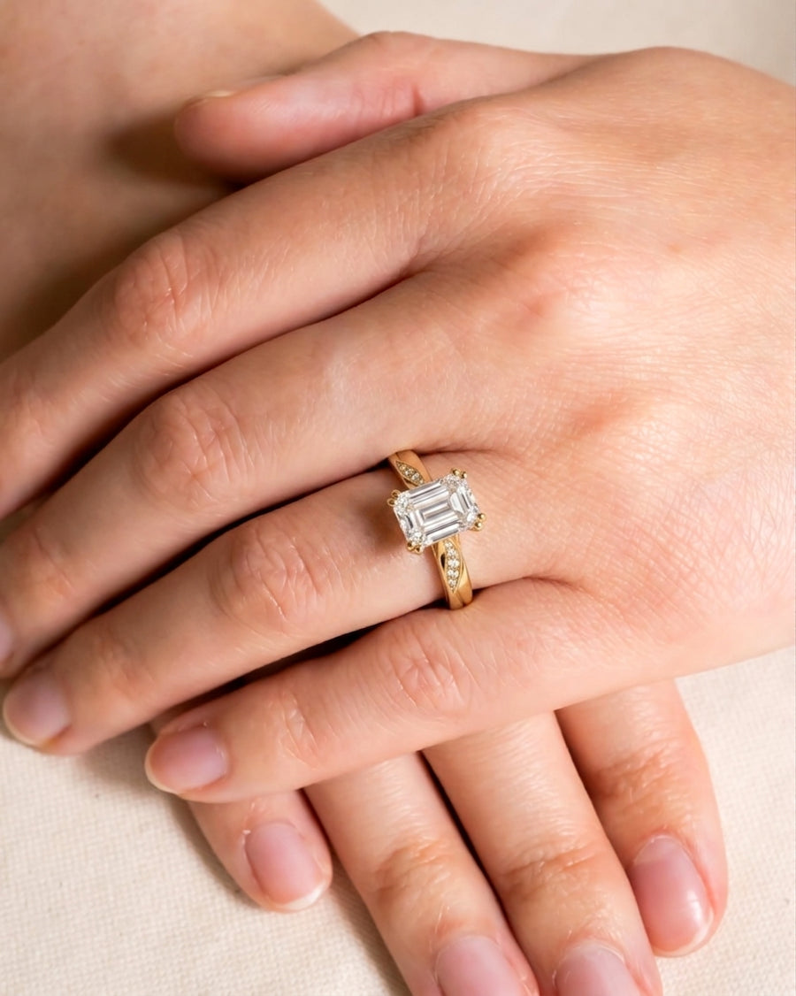 Gold engagement ring with an emerald cut diamond and pavé accents on a hand.