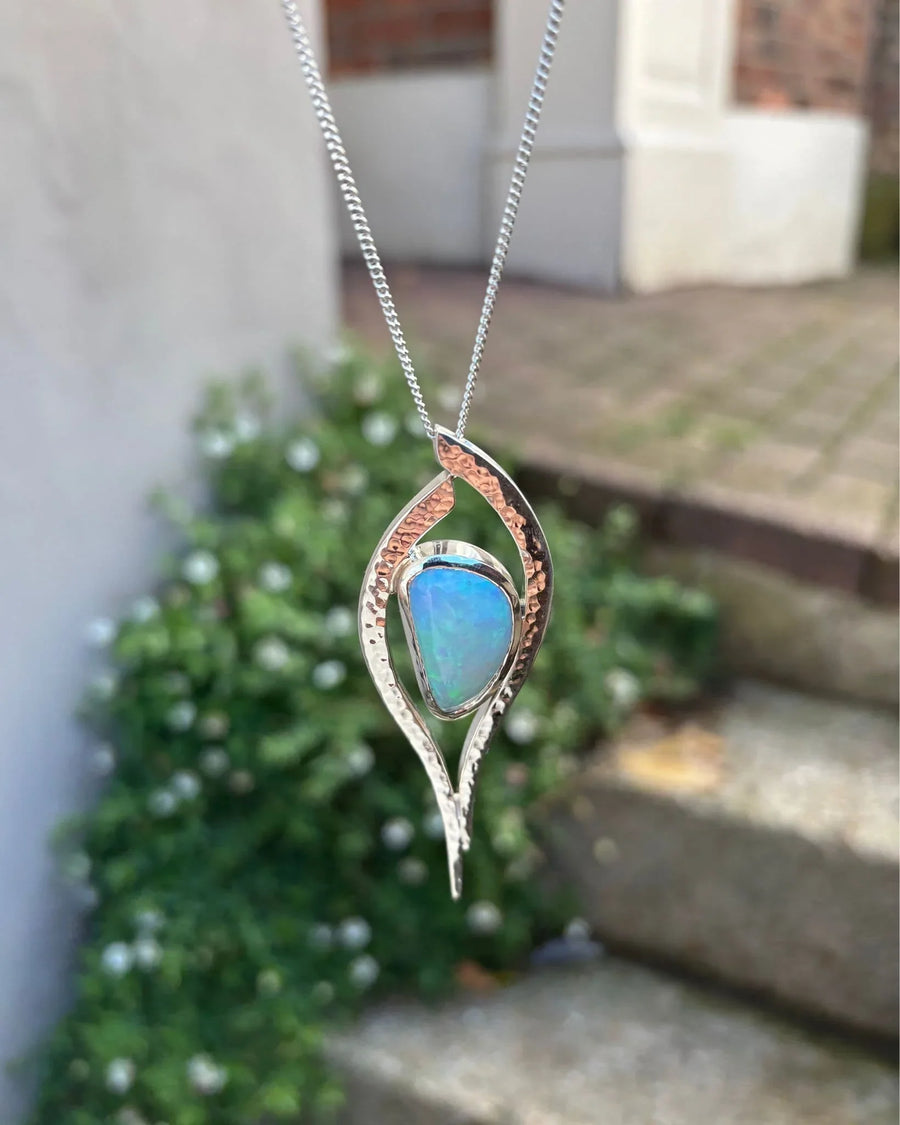 Handmade silver and gold necklace with a blue opal pendant, displayed outdoors by stone steps