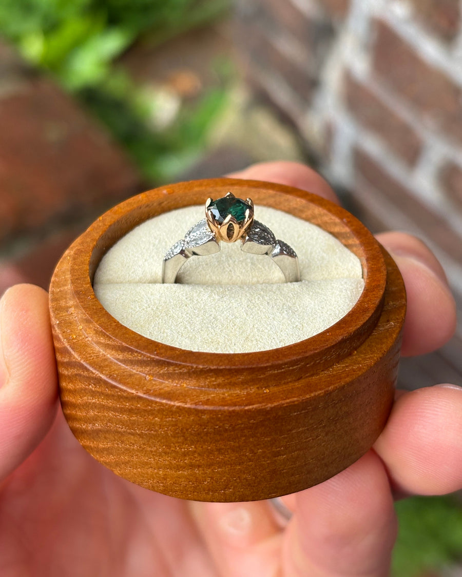 Green diamond engagement ring with a gold setting and diamond-accented silver band in a wooden box.