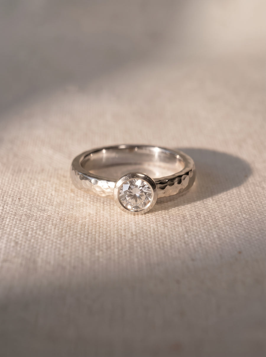 Platinum ring with a hammered finish and a bezel-set round brilliant cut diamond on a textured fabric surface.