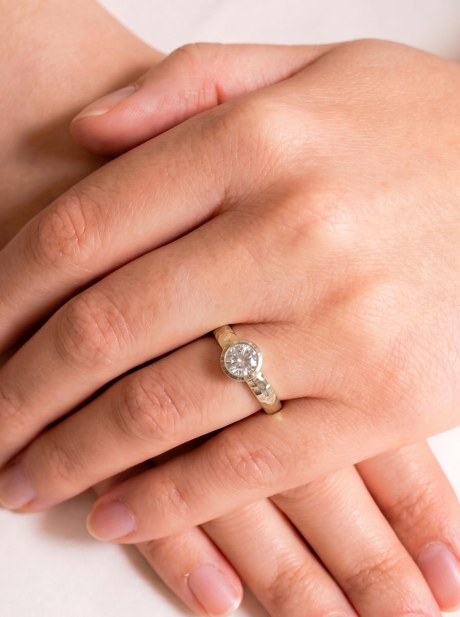 Round brilliant cut diamond ring in a hammered platinum band worn on a finger.