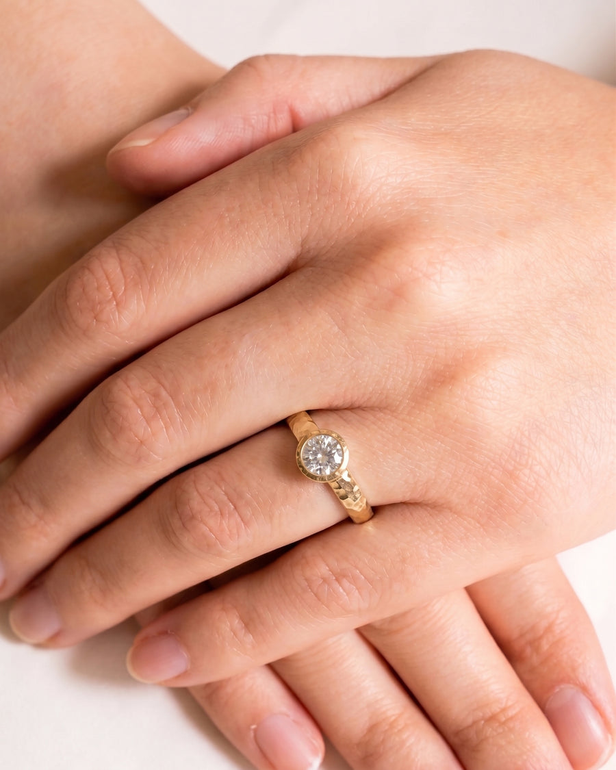 Round brilliant cut diamond set in a hammered yellow gold band on a person's finger.