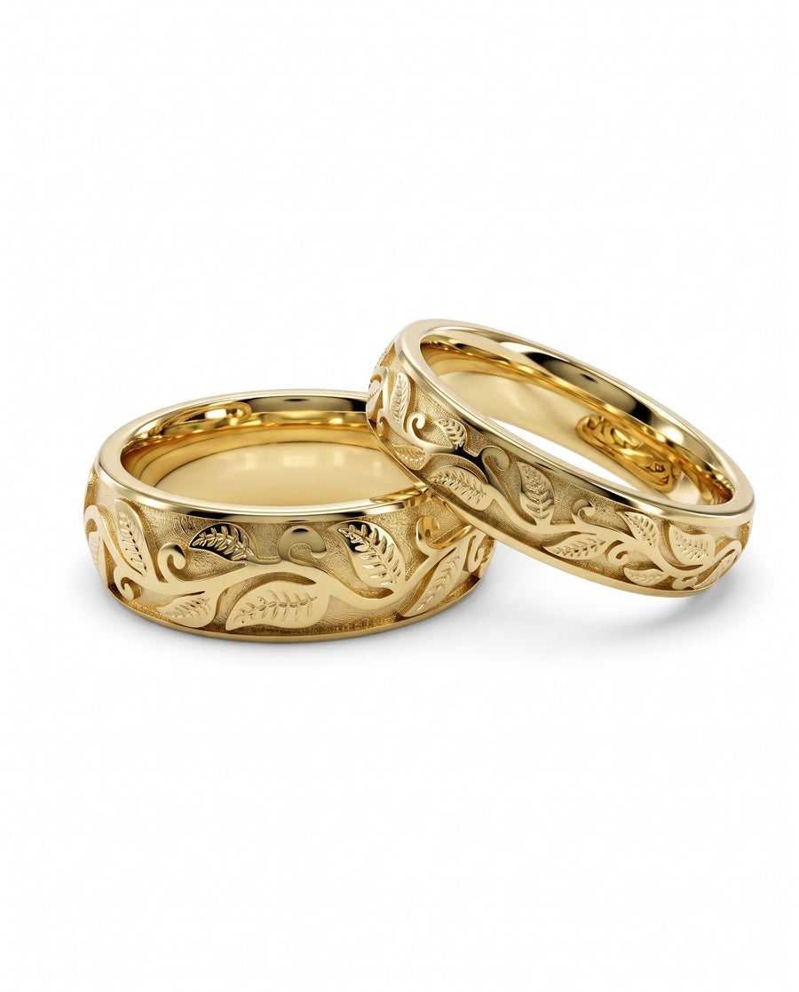 Two gold wedding bands featuring an intricate embossed leaf and vine motif against a textured background.